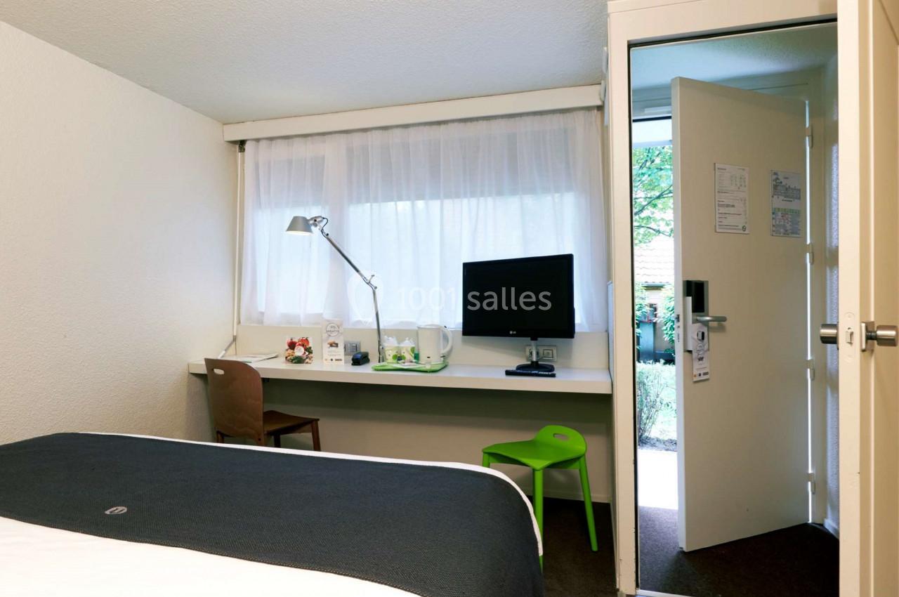 Chambre d'hôtel lumineuse avec lit, bureau équipé d'une lampe et d'un écran, et porte ouverte sur l'extérieur.