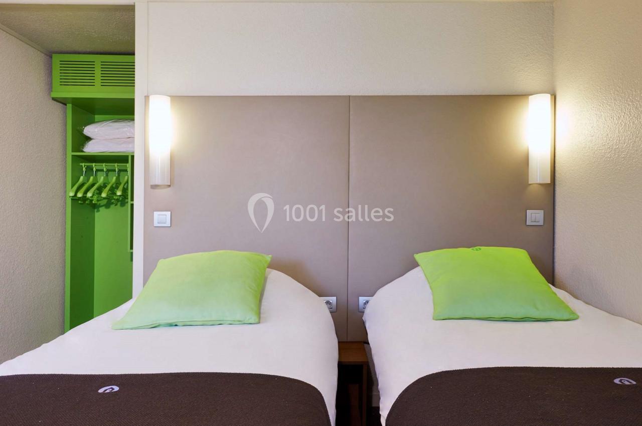 Deux lits simples avec tête de lit beige, éclairage mural et armoire ouverte avec cintres verts.