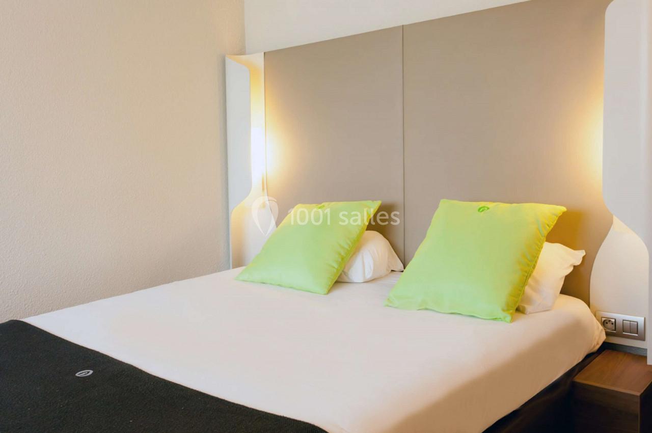 Lit double avec tête de lit rembourrée, deux coussins verts et éclairage mural intégré dans une chambre lumineuse.