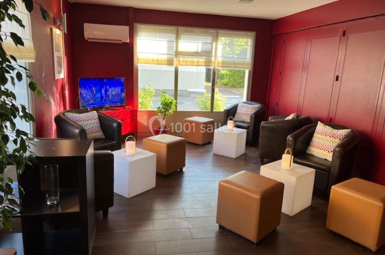Salon lumineux avec fauteuils en cuir, poufs, tables basses blanches, plantes et télévision allumée sur un mur rouge.
