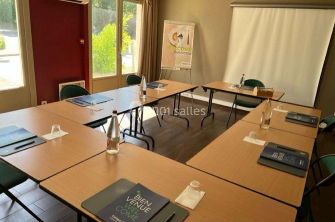 Salle de réunion lumineuse avec tables disposées en U, chaises, bloc-notes, bouteilles d'eau et écran de projection.
