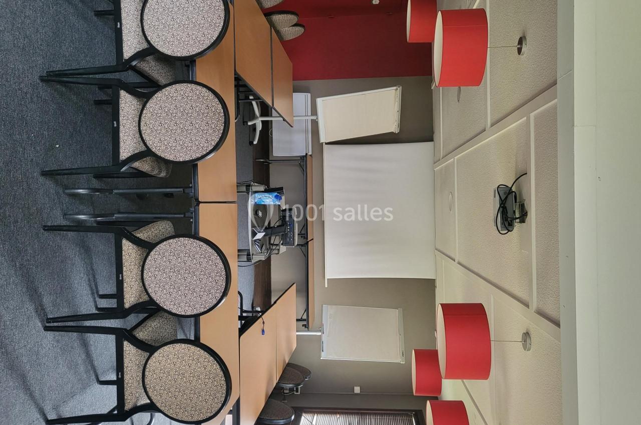 Salle de réunion avec tables en bois, chaises noires, écran de projection, paperboards et suspensions rouges.