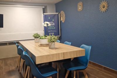 Salle de réunion lumineuse avec table en bois, chaises bleues, écran mural et affichage d'accueil.