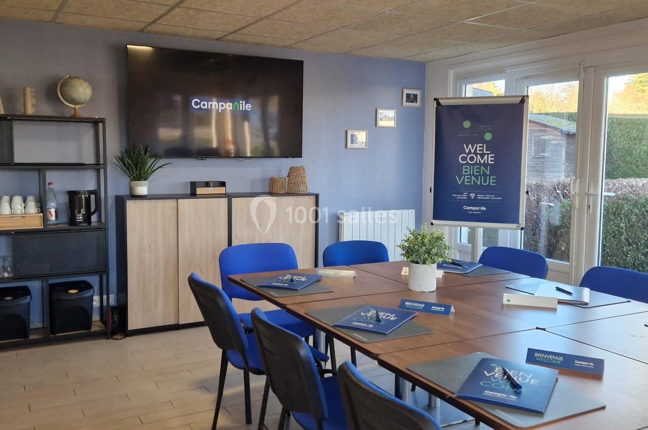 Salle de réunion lumineuse avec table, chaises bleues, écran mural, étagère et matériel de présentation.