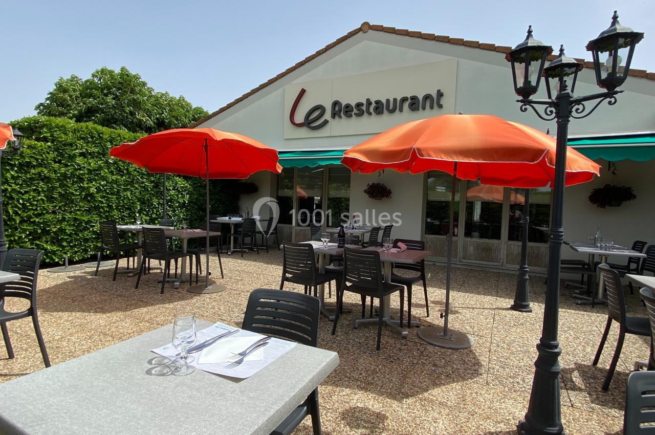 Terrasse d'un restaurant avec tables, chaises noires et parasols rouges, entourée de verdure par une journée ensoleillée.