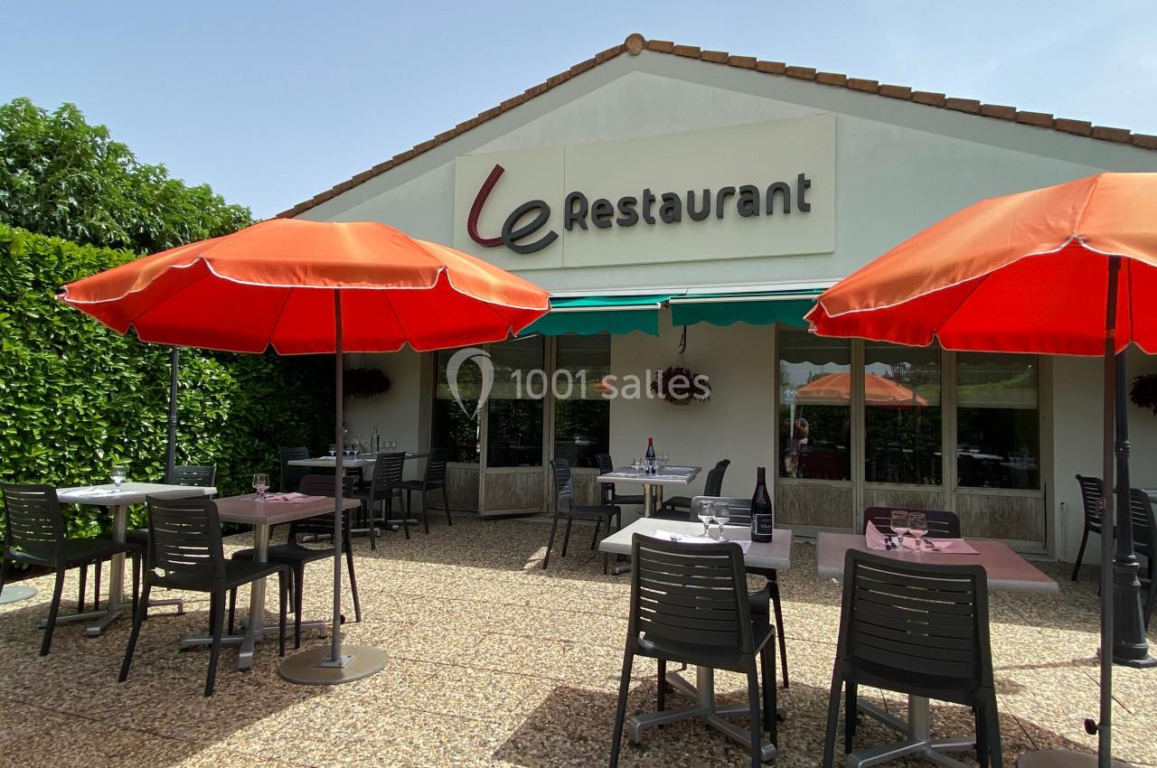 Terrasse d'un restaurant avec tables, chaises noires et parasols rouges, entourée de verdure sous un ciel dégagé.