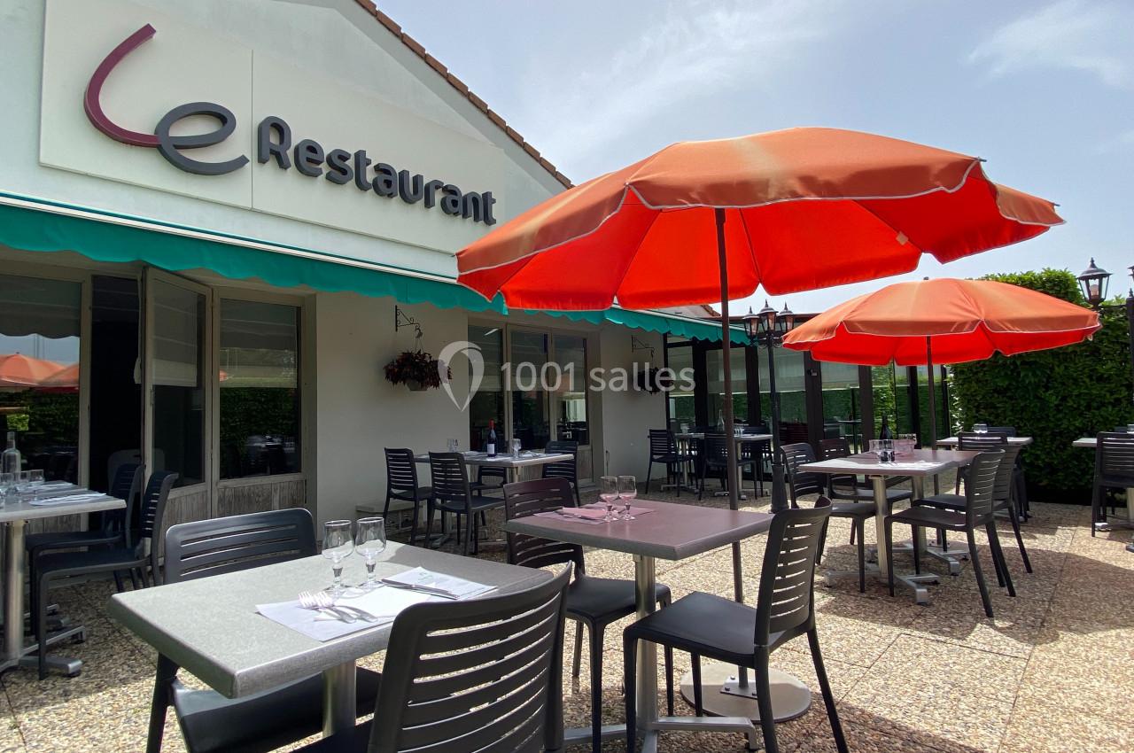 Terrasse d'un restaurant avec tables dressées, chaises noires et parasols rouges, devant une façade blanche.