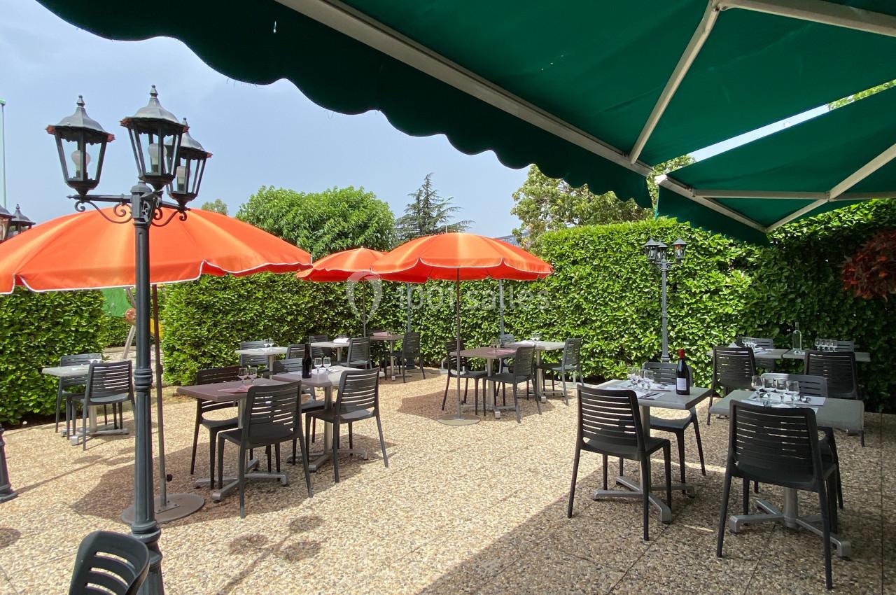Terrasse extérieure avec tables, chaises noires, parasols orange et haies vertes sous un auvent vert.