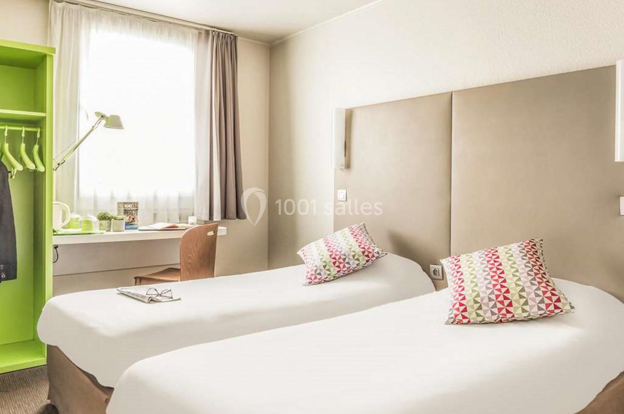 Chambre lumineuse avec deux lits simples, un bureau près de la fenêtre et une armoire ouverte avec cintres.