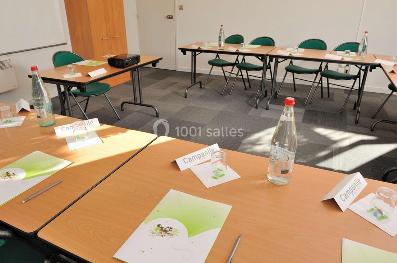 Salle de réunion lumineuse avec tables en U, chaises vertes, documents, bouteilles d'eau et matériel de présentation.