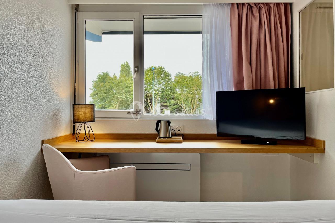 Bureau en bois avec chaise, lampe, télévision et bouilloire, placé devant une fenêtre avec vue sur des arbres.