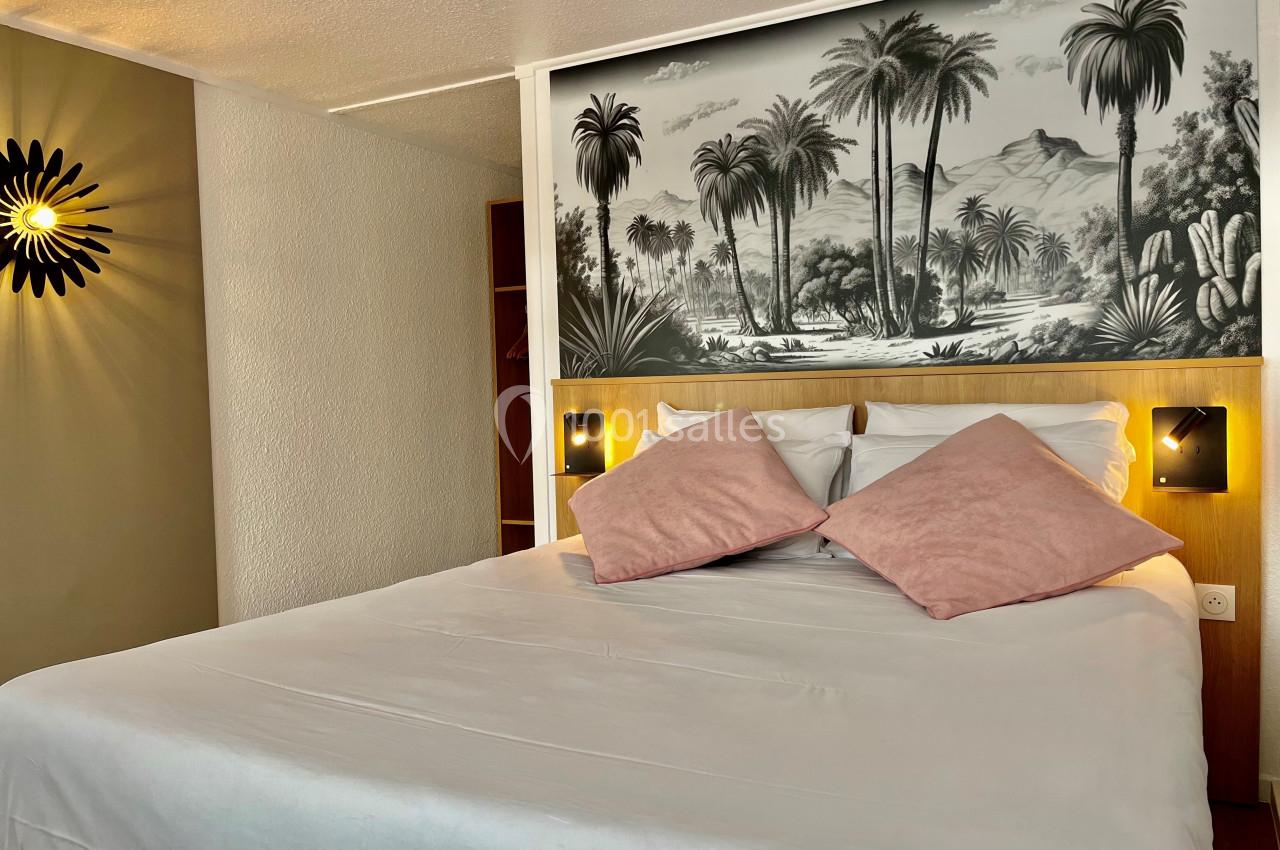 Lit double avec coussins roses, éclairé par des appliques murales, devant une fresque en noir et blanc de paysage tropical.