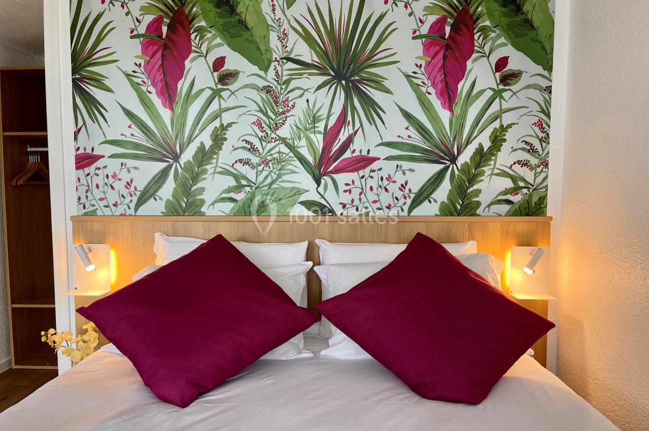 Lit double avec coussins fuchsia, éclairage d'appoint et tête de lit en bois, devant un papier peint à motifs tropicaux.