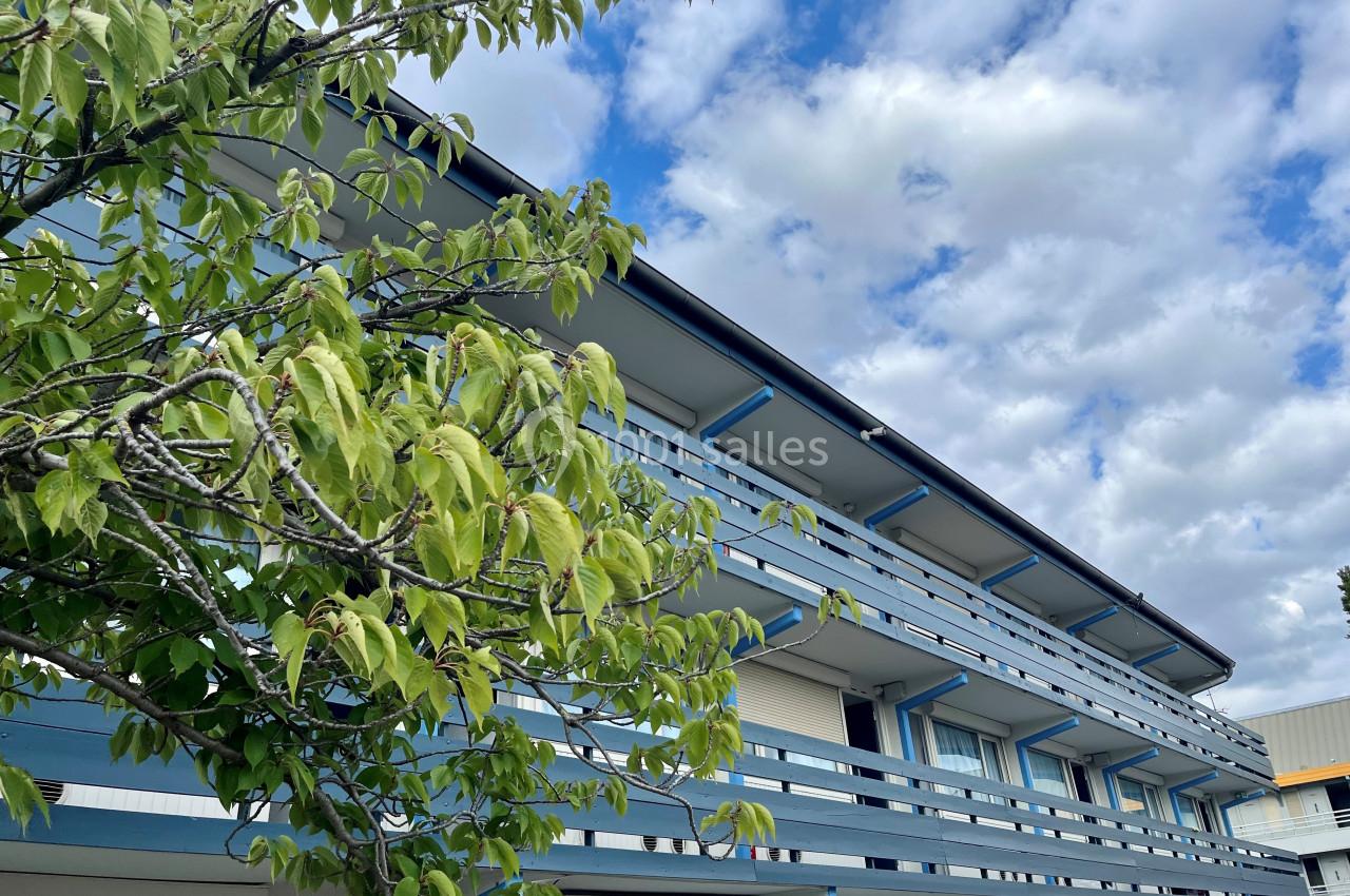 Façade d'un bâtiment à étages avec balcons bleus, partiellement cachée par un arbre sous un ciel nuageux.
