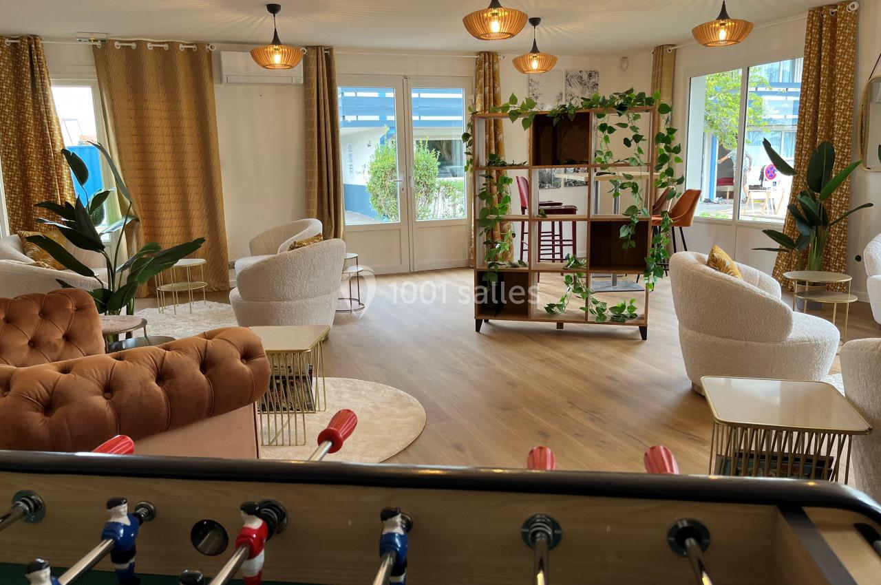 Espace lumineux avec fauteuils, étagères décorées, baby-foot au premier plan et grandes fenêtres donnant sur l'extérieur.