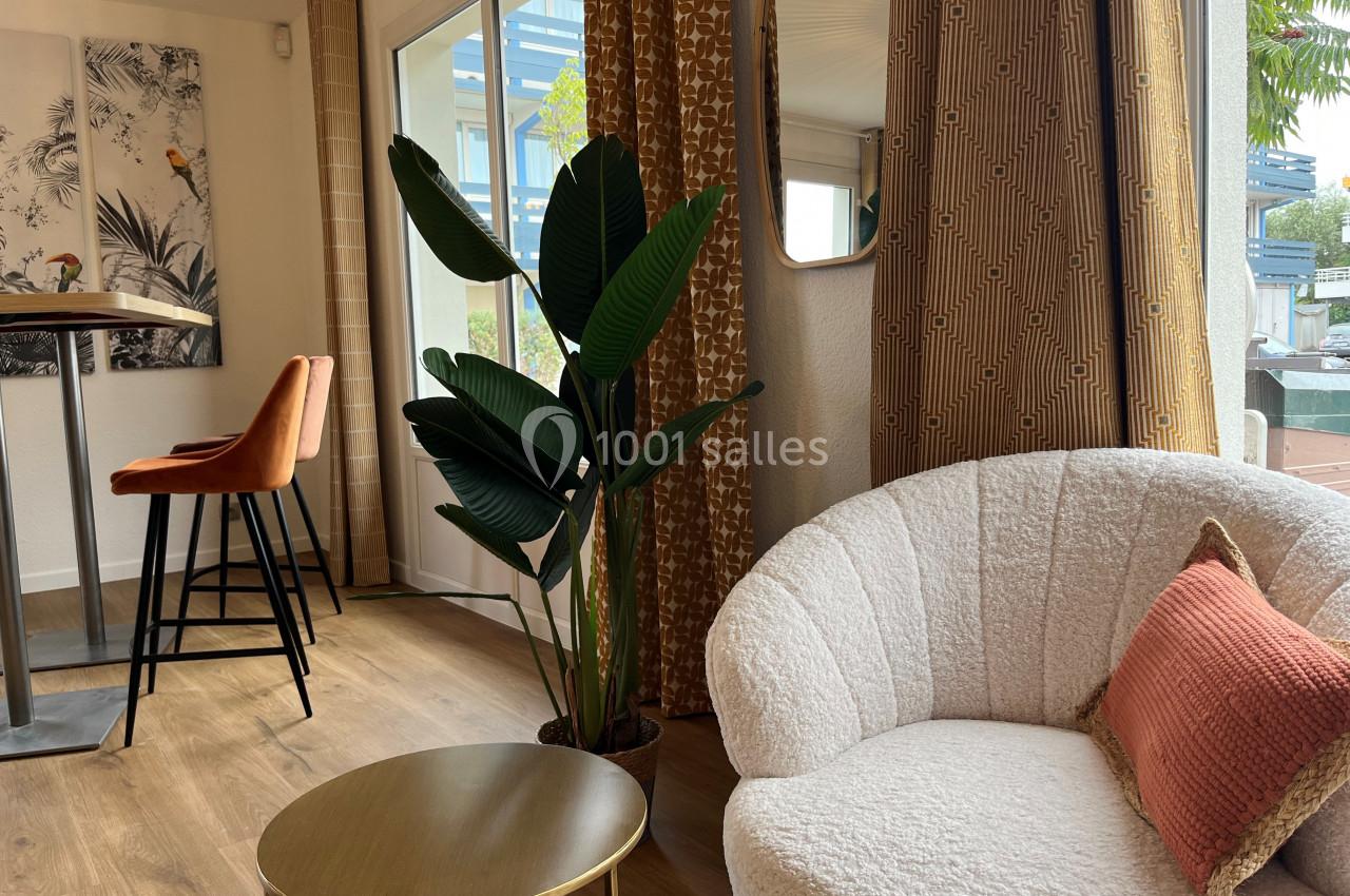 Coin salon lumineux avec fauteuil blanc, table basse dorée, plante verte et tabourets près d'une table haute.