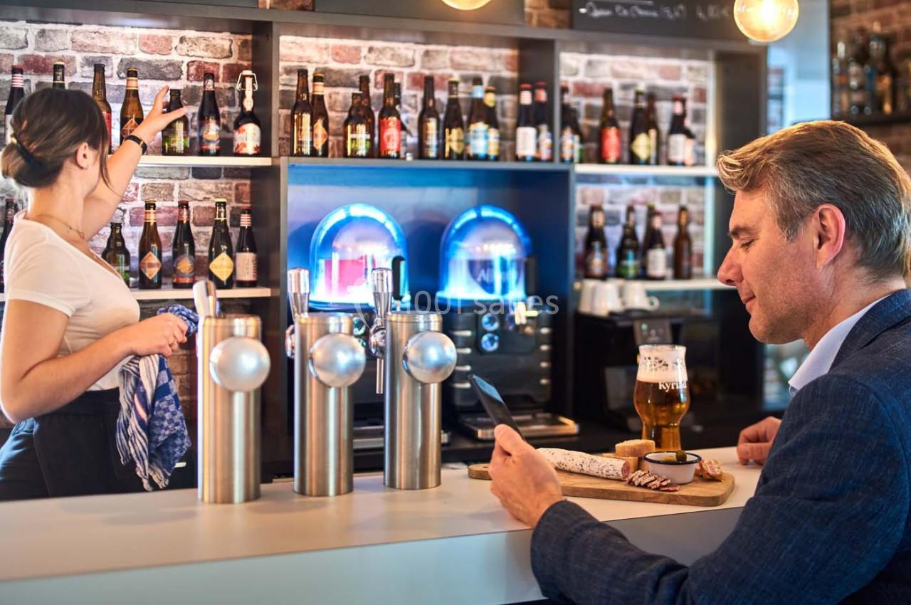 Un homme au comptoir d'un bar lit sur son téléphone tandis qu'une serveuse range des bouteilles sur une étagère.