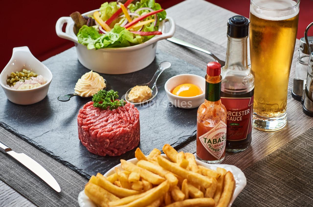 Assiette de tartare de bœuf cru avec frites, salade, condiments variés et verre de bière sur une table en bois.