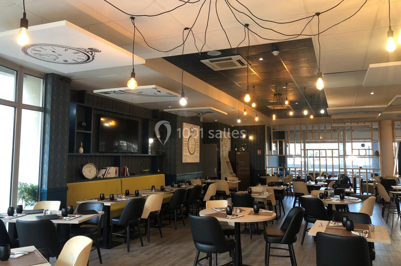 Salle de restaurant lumineuse avec tables dressées, chaises noires et décoration murale sur le thème des horloges.