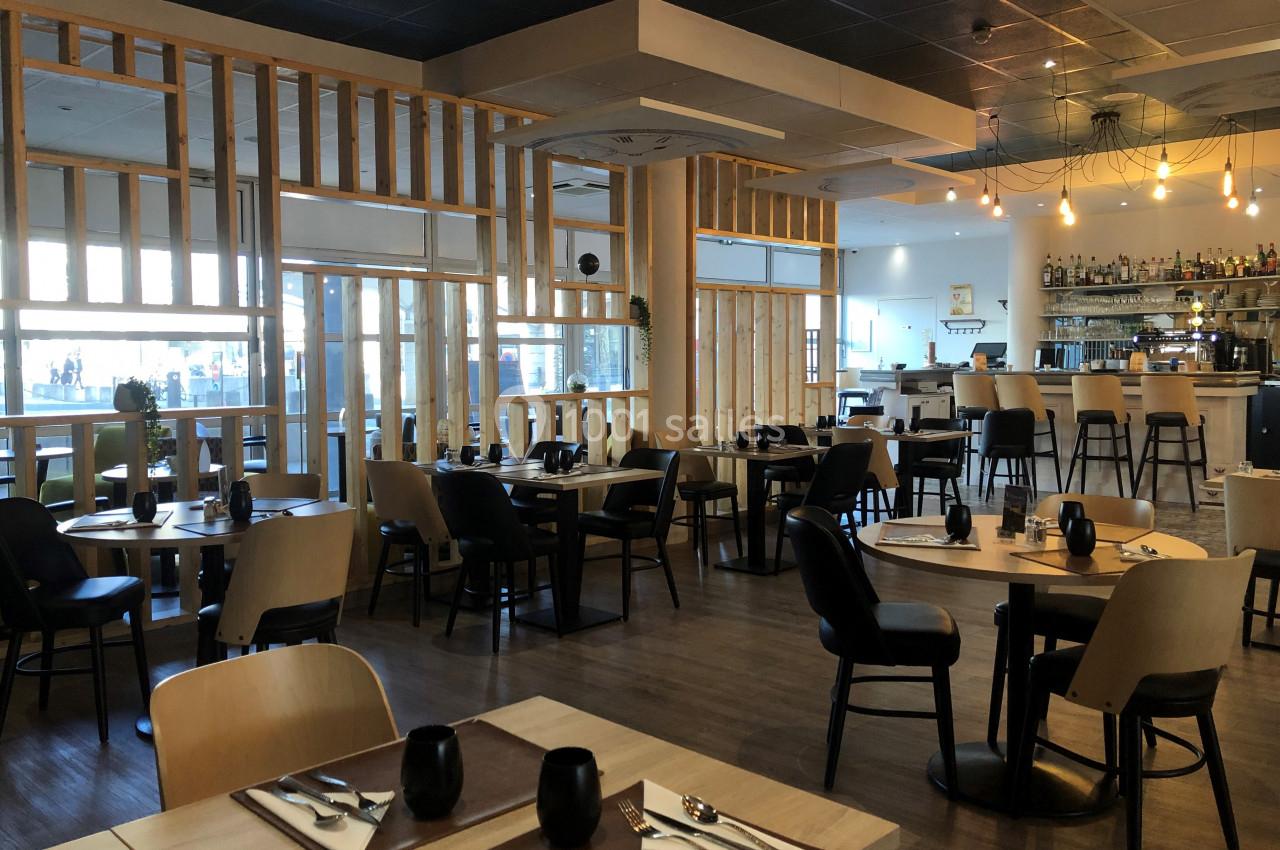 Salle de restaurant moderne avec tables dressées, chaises en bois, bar en arrière-plan et éclairage suspendu.