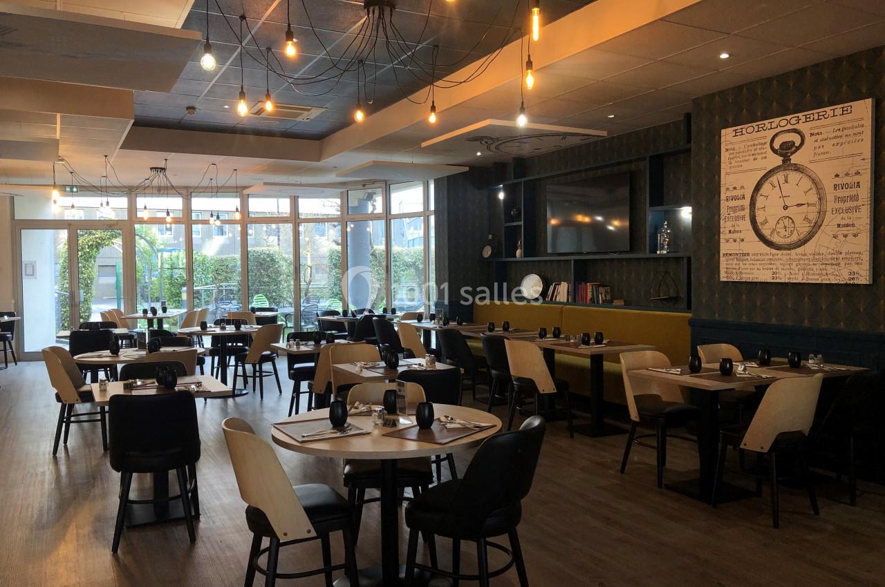 Salle de restaurant lumineuse avec tables dressées, décorée de luminaires suspendus et d'une grande horloge murale.