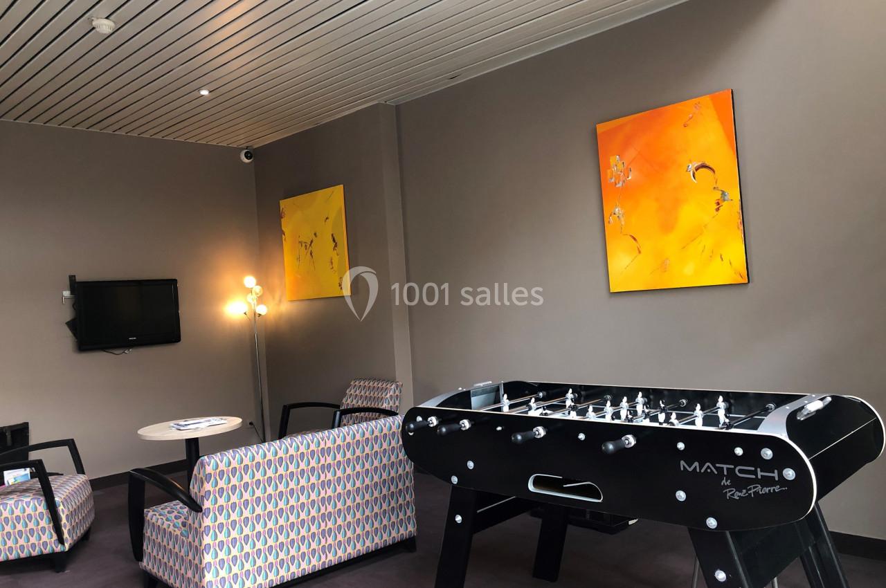 Salle de détente avec baby-foot, fauteuils, table basse, télévision murale et tableaux colorés accrochés au mur.