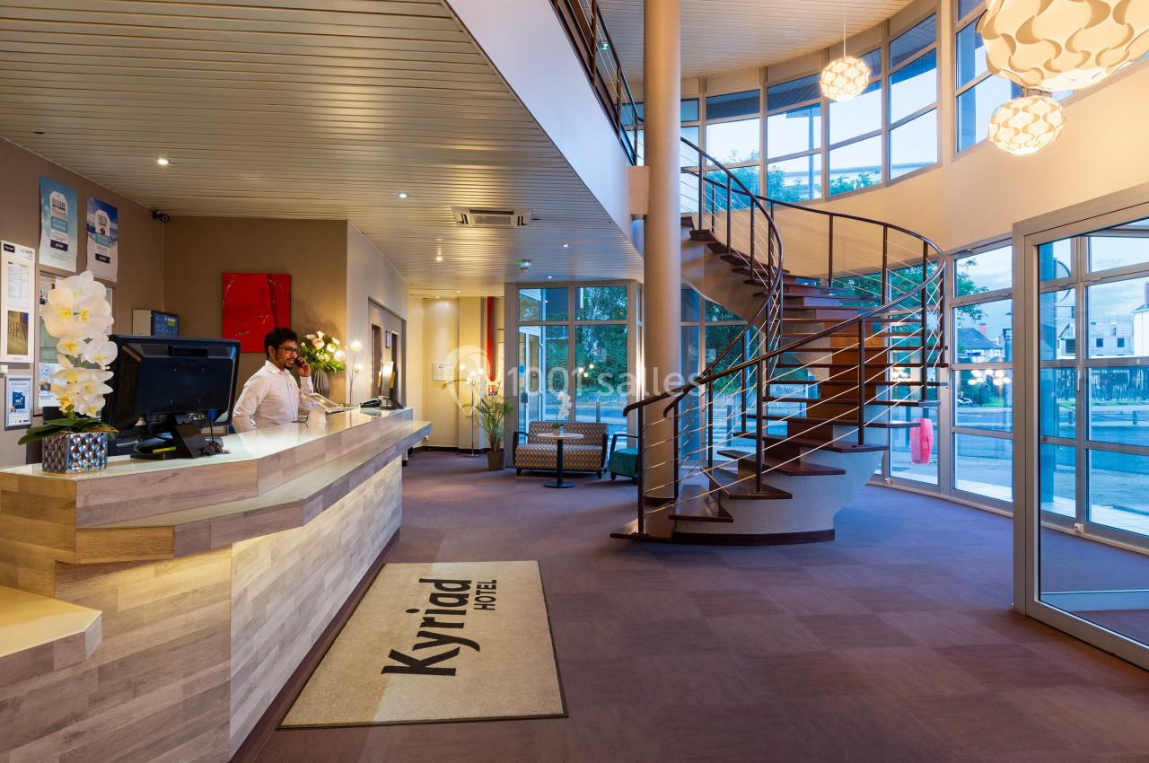 Réception d'hôtel avec comptoir en bois, escalier en colimaçon, grandes baies vitrées et éclairage chaleureux.