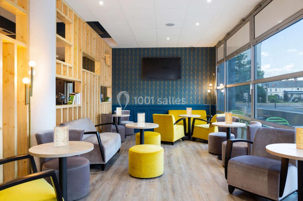 Espace lounge lumineux avec fauteuils gris et jaunes, tables rondes en bois, mur décoratif et grande baie vitrée.