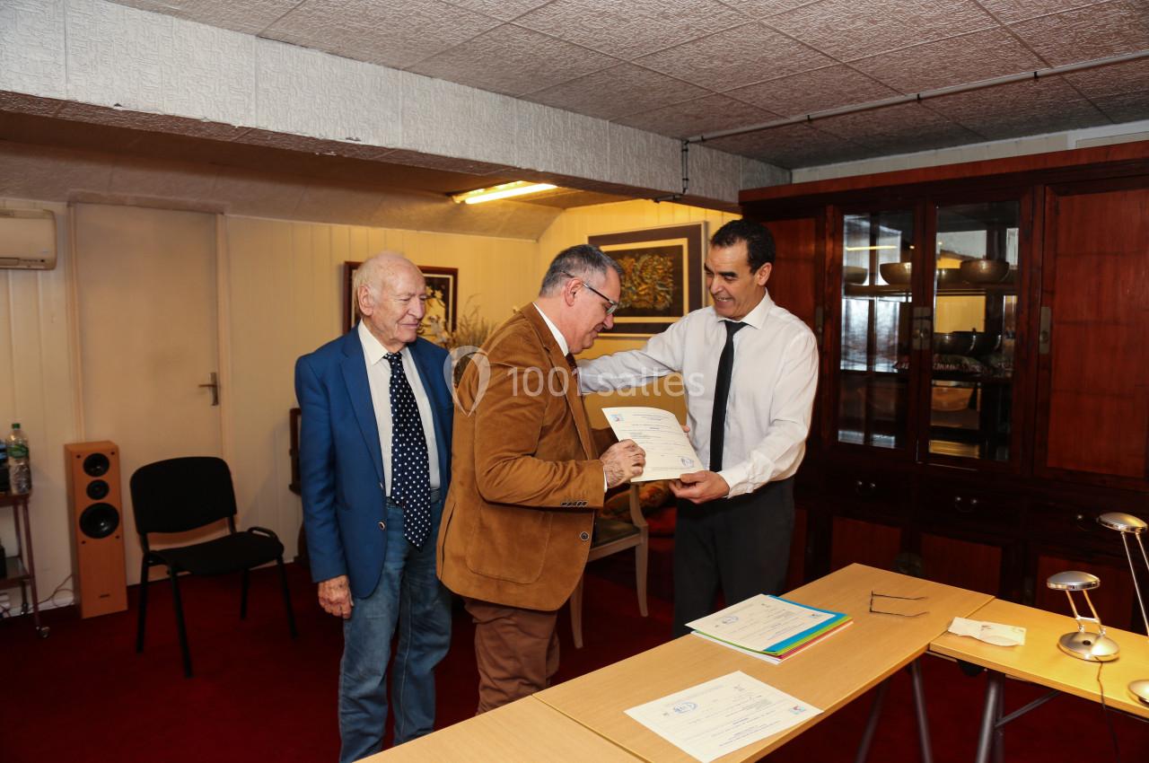 Un homme remet un certificat à un autre homme dans une pièce avec des meubles en bois et un tapis rouge.
