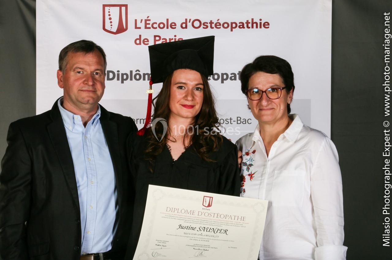 Une diplômée en tenue de cérémonie tient son diplôme, entourée de deux personnes devant une bannière d'école d'ostéopathie.