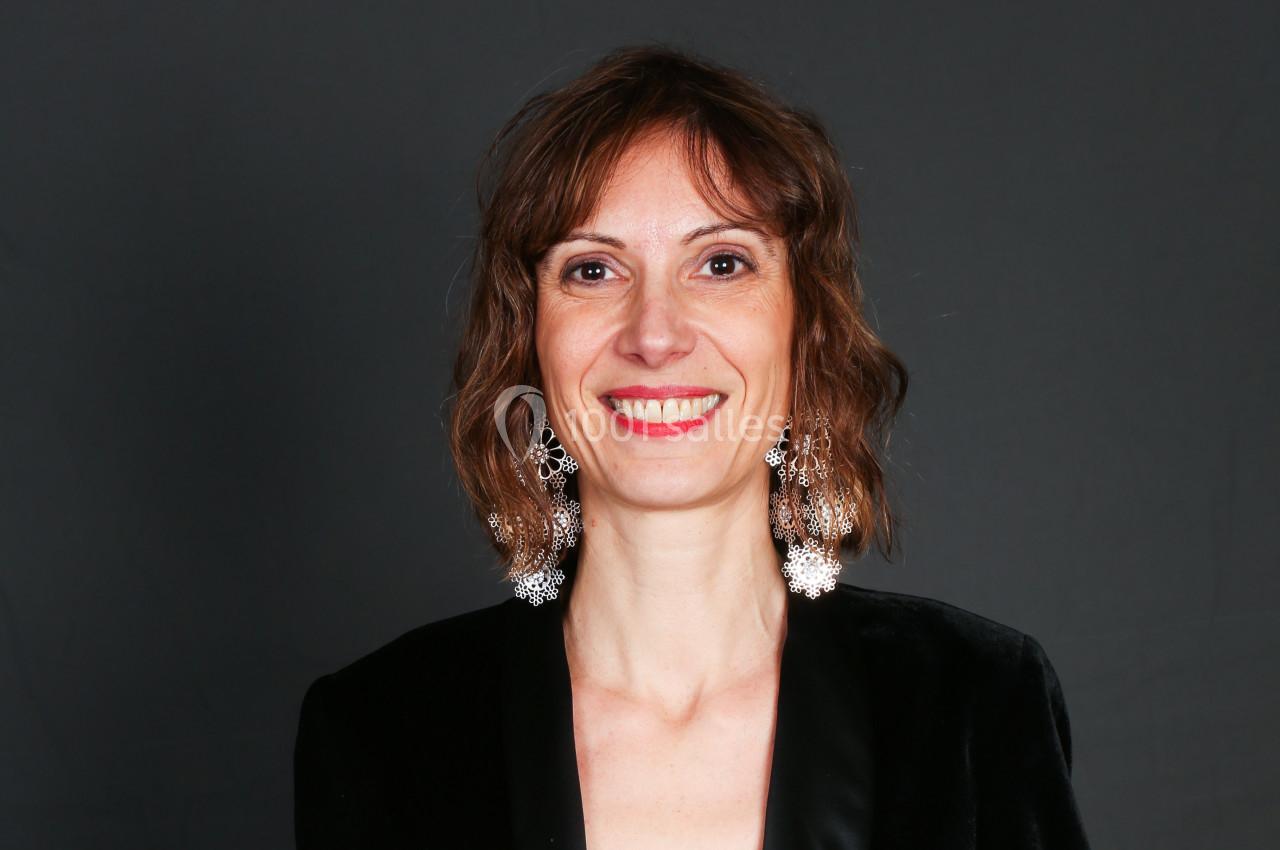 Femme souriante portant des boucles d'oreilles pendantes et une veste noire, sur fond gris uni.