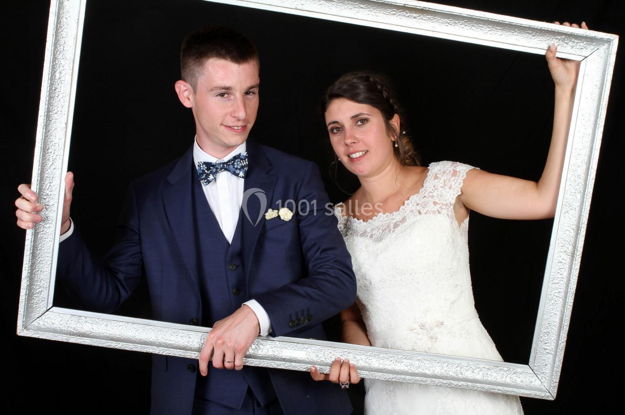 Un couple en tenue de mariage pose avec un grand cadre argenté sur un fond noir.