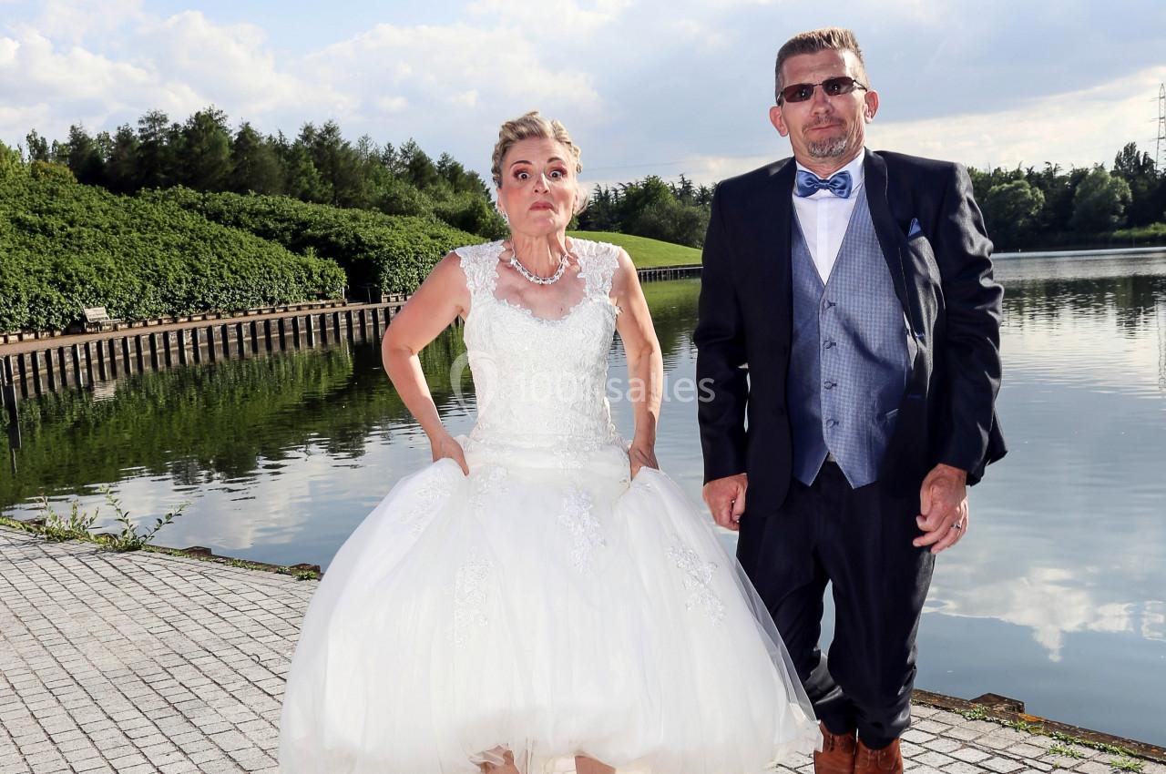 Une femme en robe de mariée et un homme en costume posent debout près d'un plan d'eau bordé de végétation.