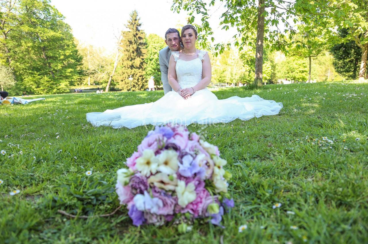 Un couple en tenue de mariage assis sur l'herbe dans un parc, avec un bouquet flou au premier plan.