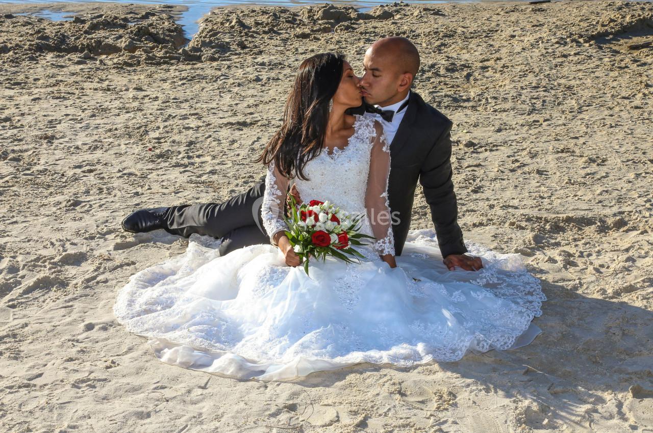 Un couple en tenue de mariage s'embrasse assis sur le sable près de l'eau, la mariée tenant un bouquet de fleurs.