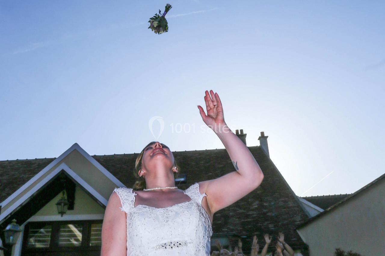 Une mariée lance son bouquet en l'air devant un bâtiment, des mains tendues en arrière-plan.
