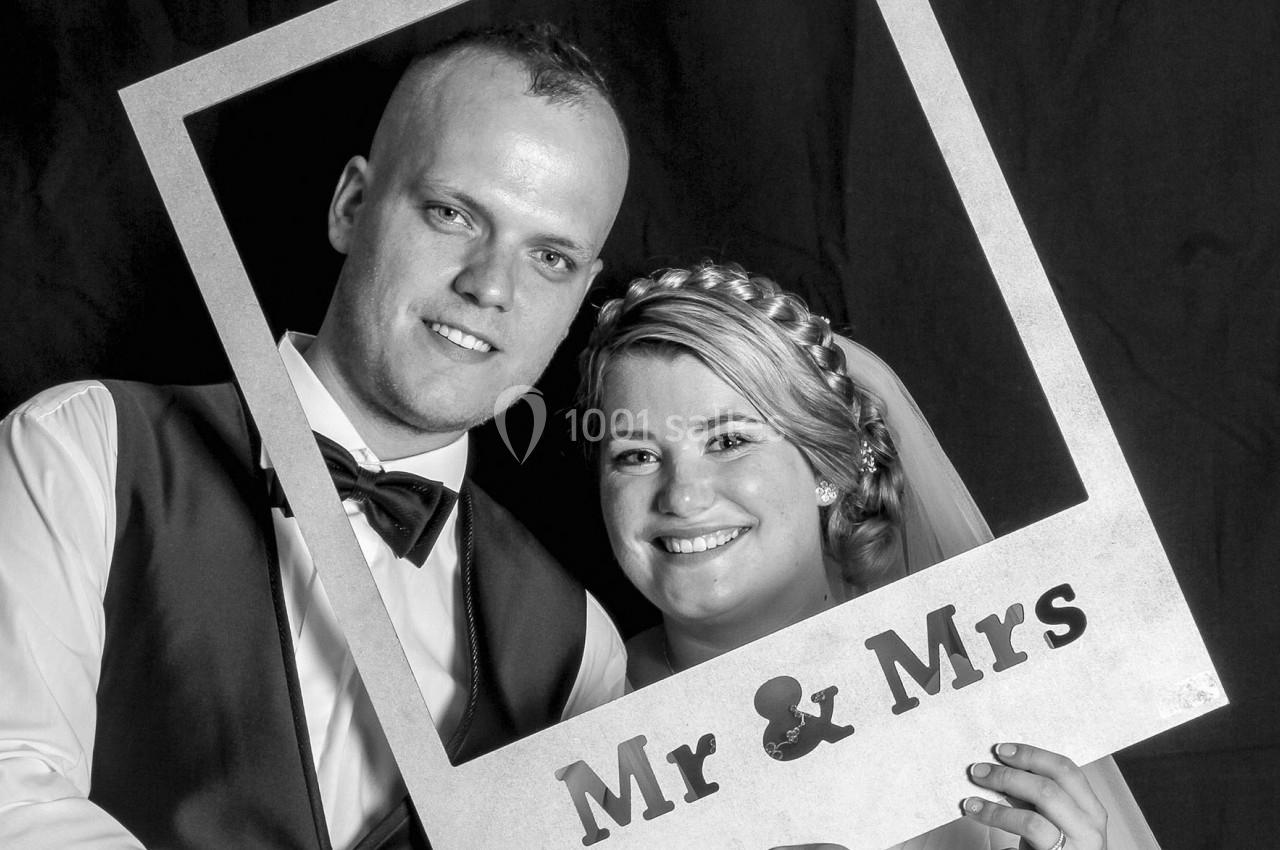 Un couple souriant pose avec un cadre photo portant l'inscription ’Mr & Mrs’ sur un fond sombre.