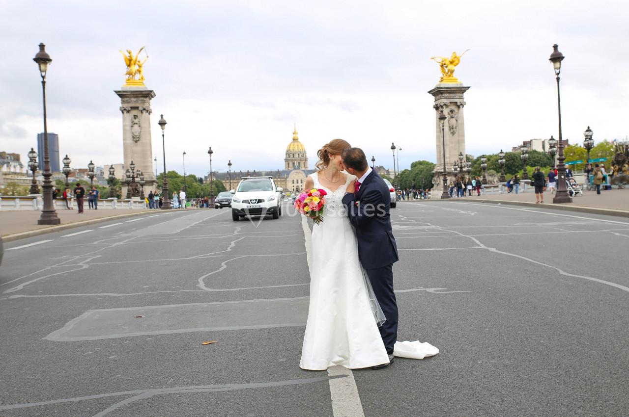 Un couple en tenue de mariage pose sur un pont parisien avec des lampadaires et des statues dorées en arrière-plan.