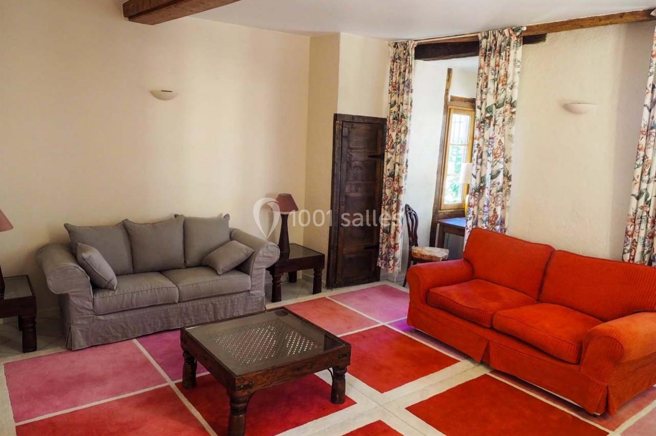 Salon avec deux canapés, table basse en bois, tapis coloré et rideaux fleuris, éclairé par une fenêtre.