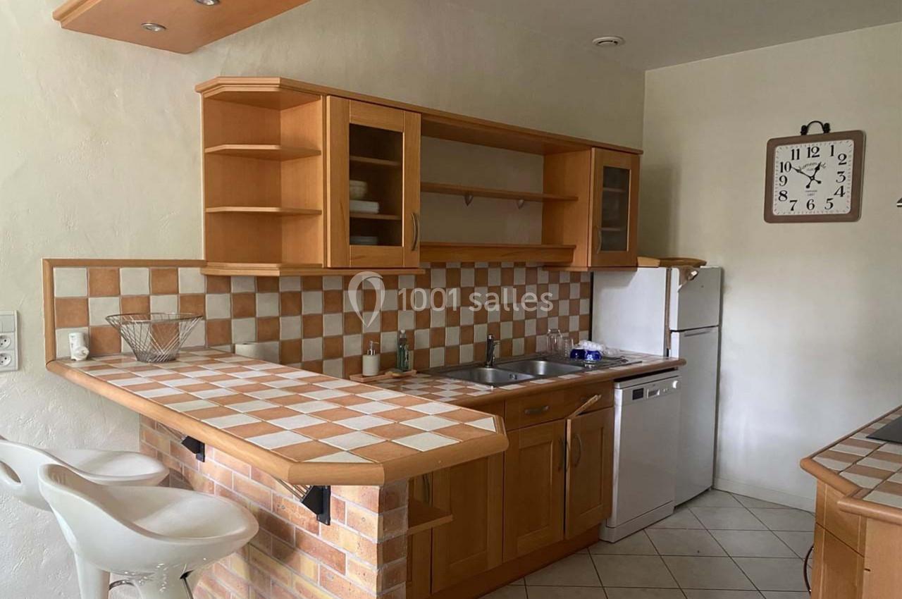 Cuisine avec meubles en bois, plan de travail carrelé, évier inox, électroménagers et horloge murale.