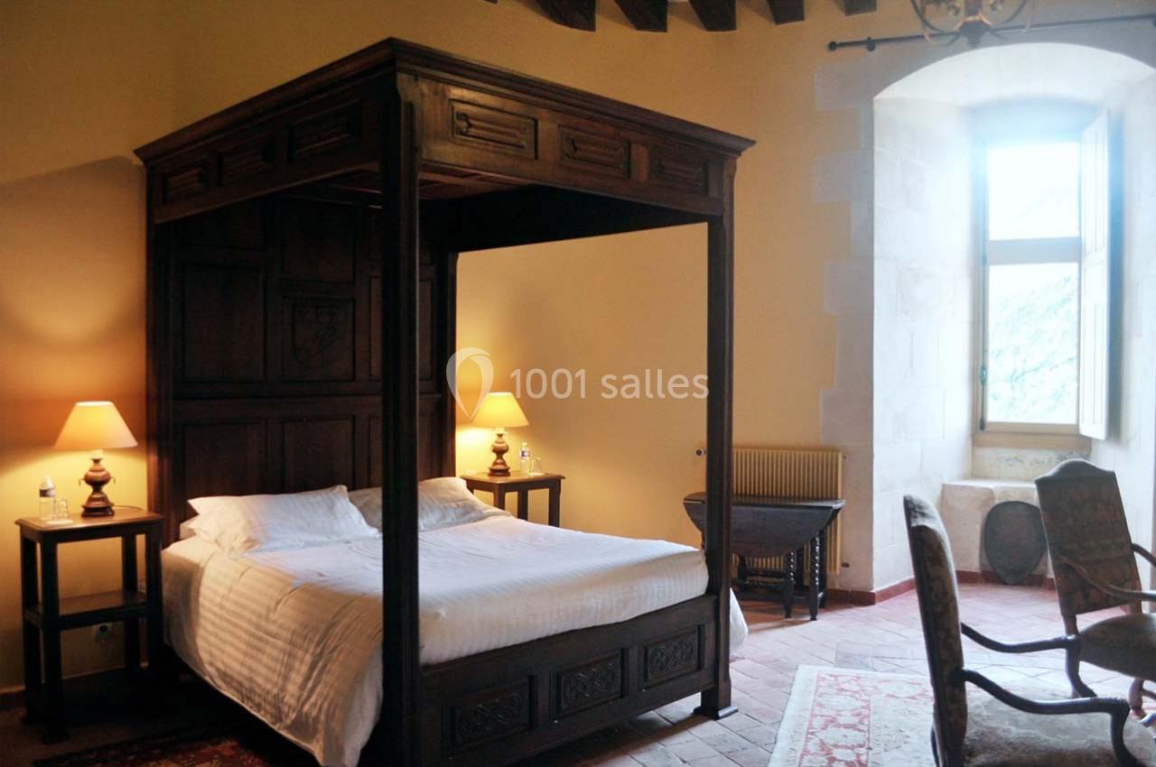 Lit à baldaquin en bois sombre dans une chambre lumineuse avec murs en pierre, fenêtres ouvertes et lampes allumées.