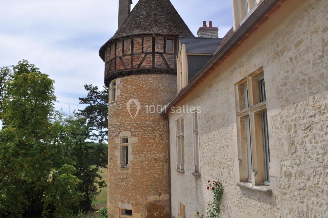 Façade d'un bâtiment ancien en pierre avec une tour ronde, entouré de verdure sous un ciel dégagé.