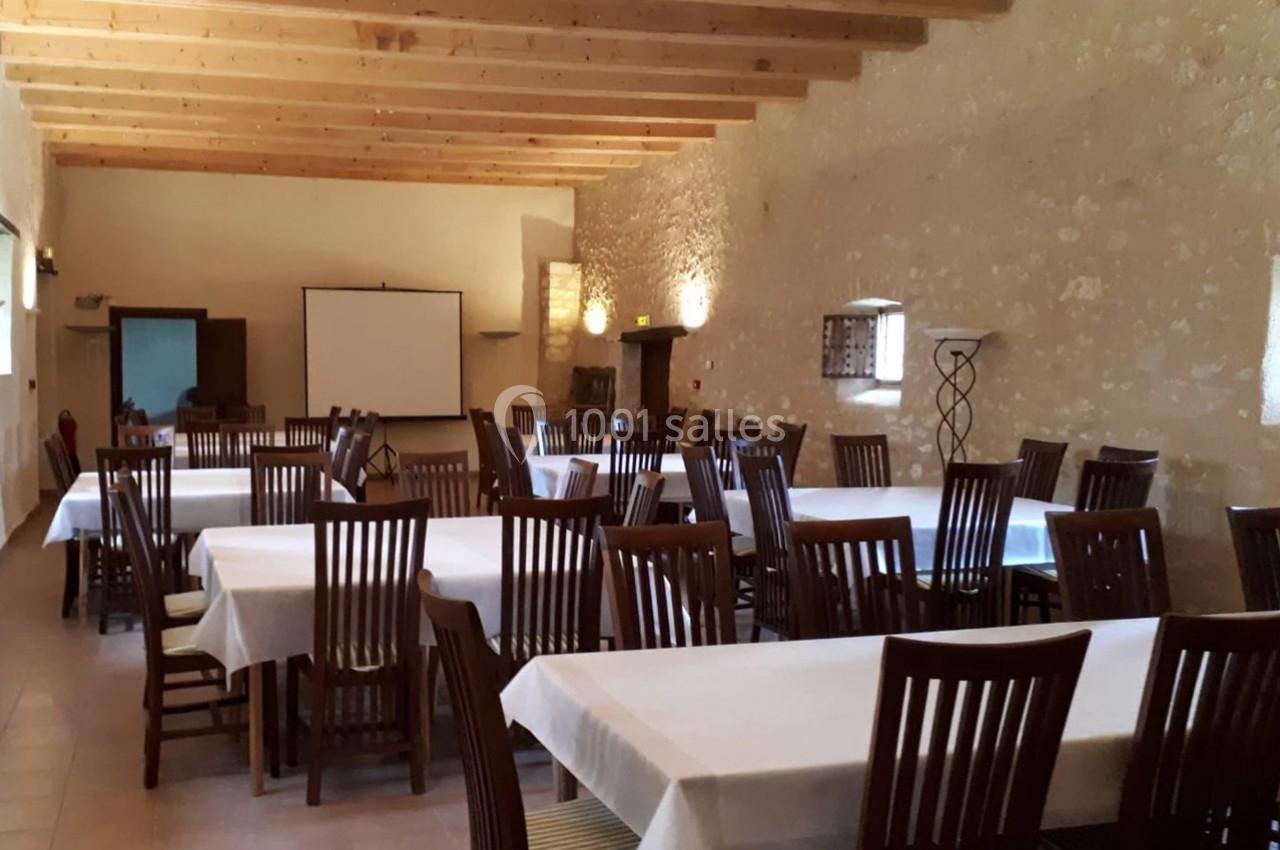 Salle avec des tables rectangulaires couvertes de nappes blanches et des chaises en bois, éclairée par des appliques murales.