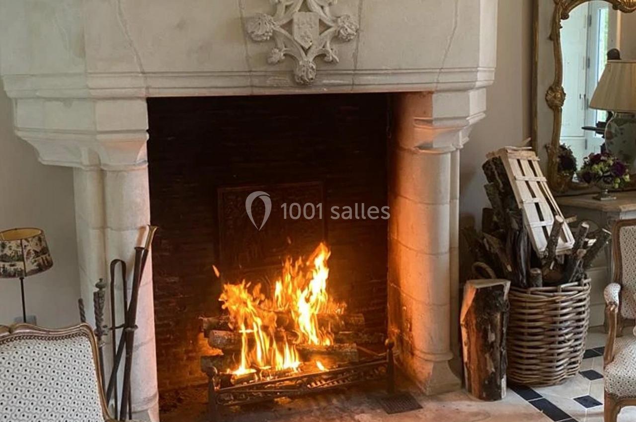 Cheminée en pierre allumée avec des bûches enflammées, entourée de fauteuils et d'un panier de bois.