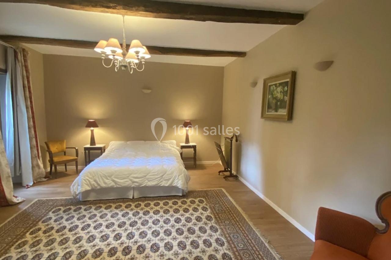 Chambre lumineuse avec lit double, tapis au sol, poutres apparentes, mobilier classique et éclairage doux.