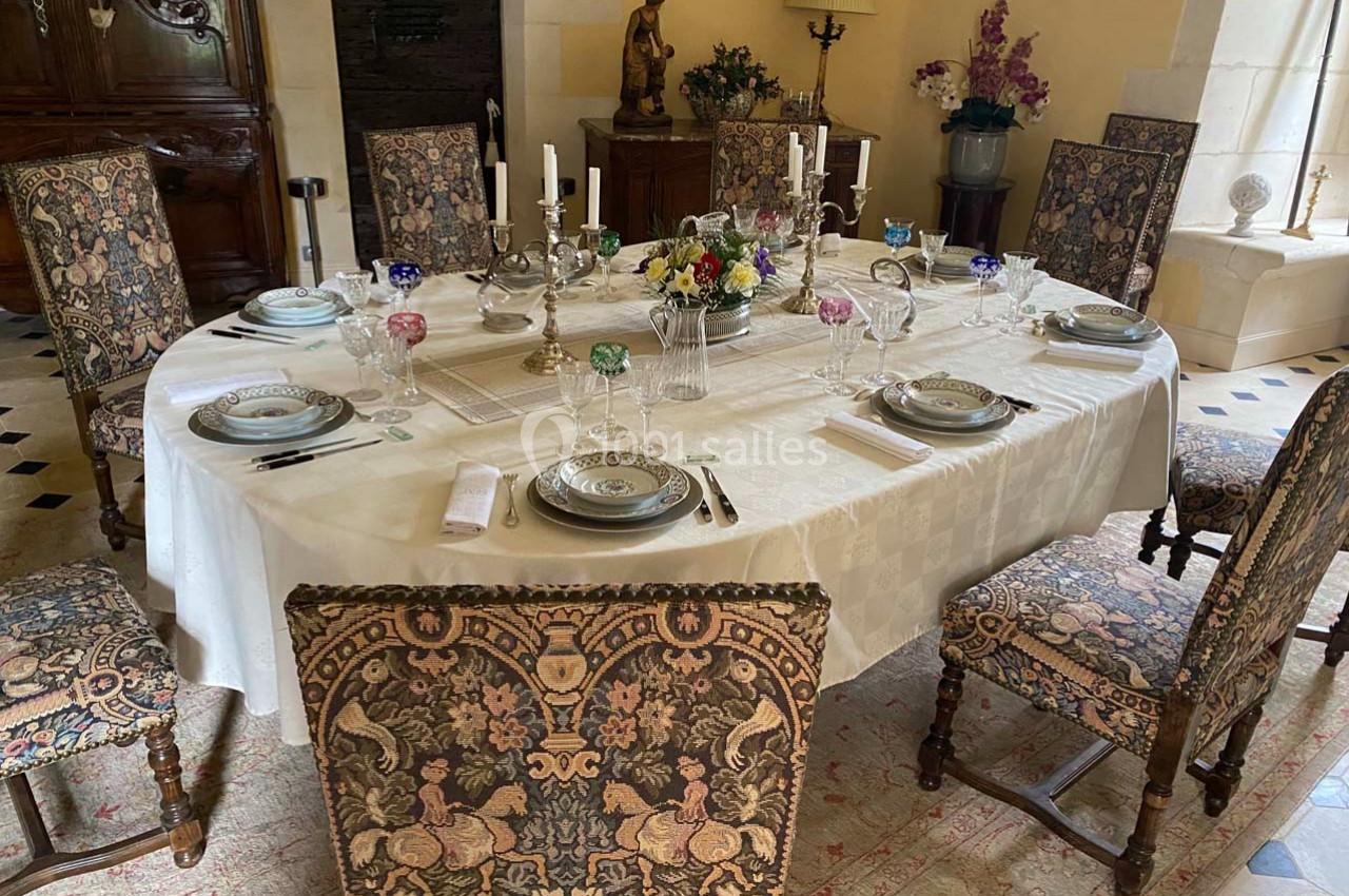 Table ovale dressée avec vaisselle, chandeliers et fleurs, entourée de chaises à motifs dans une salle élégante.