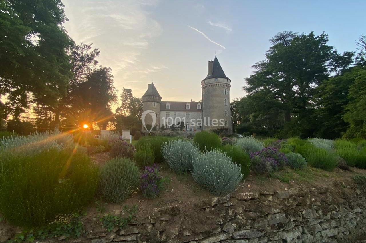Château en pierre entouré de jardins fleuris, au coucher du soleil, avec des arbres en arrière-plan.