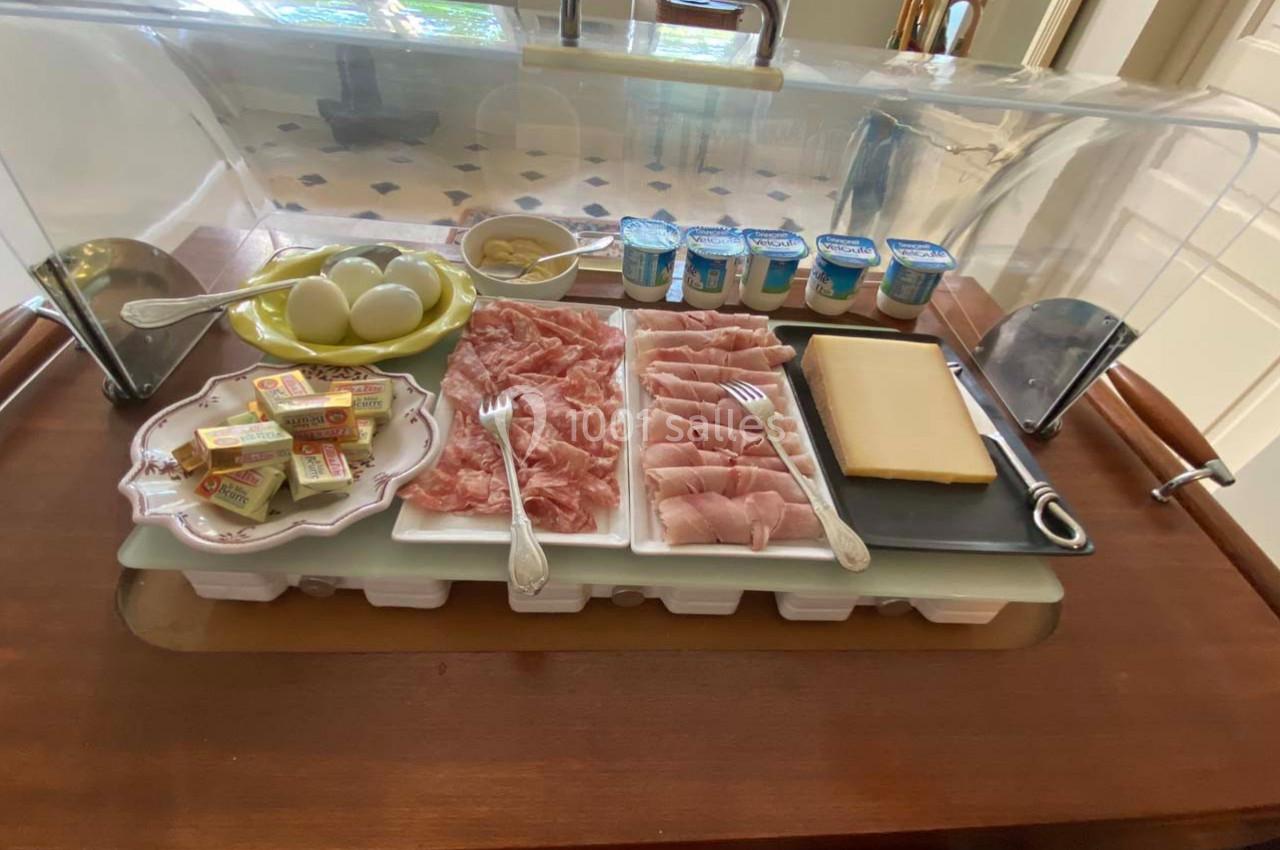 Plateau de petit-déjeuner avec charcuterie, fromage, œufs durs, beurre, yaourts et moutarde, sous une cloche transparente.