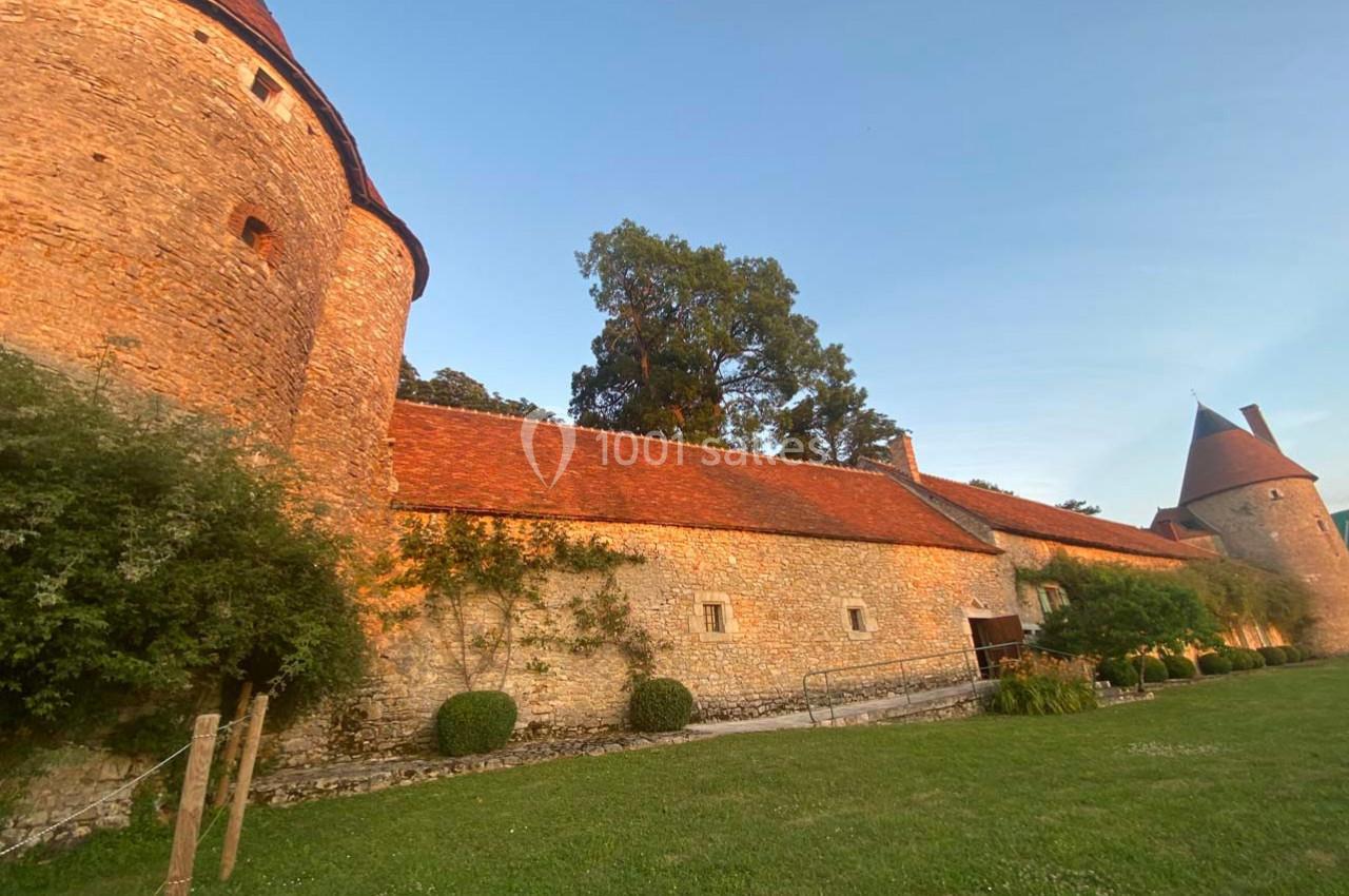 Mur en pierre d'un château avec tours rondes et toits en tuiles rouges, entouré de verdure au coucher du soleil.
