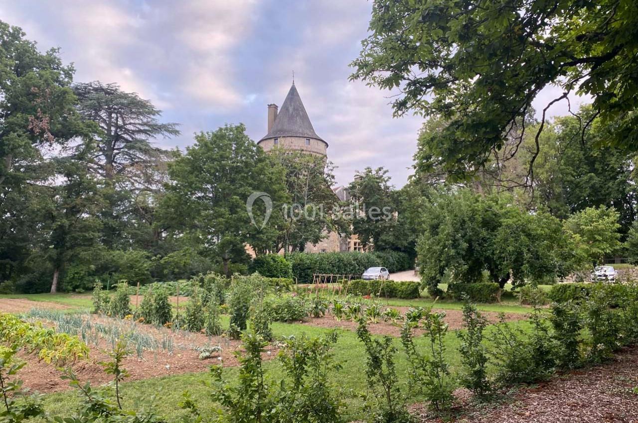 Potager verdoyant avec des rangées de légumes, entouré d'arbres, et un château en arrière-plan sous un ciel nuageux.
